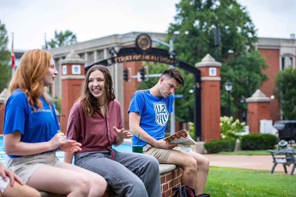 Students sit on the edge of the dolphin pond with Grand Blvd and the Saint Louis University gateway in the background on a sunny day