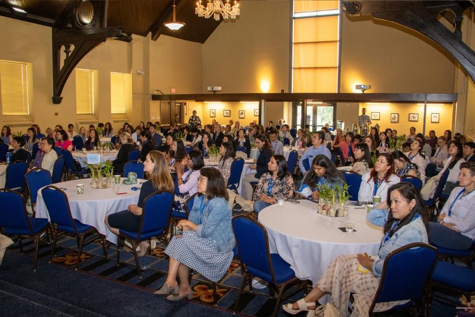 A crowded room of conference attendees are seen from the front while sitting at tables, watching a presentation